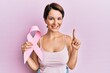 © Krakenimages.com - Young brunette woman with short hair holding pink cancer ribbon smiling with an idea or question pointing finger with happy face, number one
