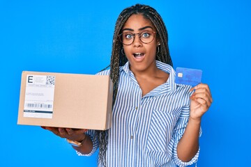 Canvas Print - Young african american woman with braids holding delivery box and credit card afraid and shocked with surprise and amazed expression, fear and excited face.