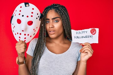 Wall Mural - Young african american woman with braids holding halloween banner and scary mask clueless and confused expression. doubt concept.