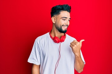 Wall Mural - Young man with beard listening to music using headphones smiling with happy face looking and pointing to the side with thumb up.