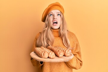 Wall Mural - Beautiful young caucasian girl wearing french look with beret holding baguettes bread angry and mad screaming frustrated and furious, shouting with anger looking up.