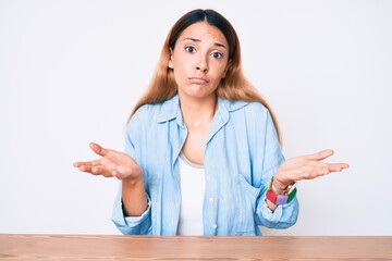 Sticker - Young brunette woman sitting on the table wearing casual clothes clueless and confused expression with arms and hands raised. doubt concept.