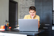 © Natali - Cute young teenager in yellow shirt sitting behind desk in kitchen next to laptop and study. Serious boy makes homework, listening lesson. Home, distance education, self study by kids.