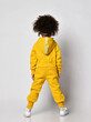 Curly dark-skinned kid girl in warm blue sports jumpsuit with hood and sneakers stands back to camera over white wall background, rear view. Trendy children fashion, stylish outfit