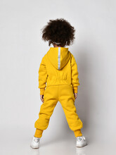 Curly Mulatto Dark-skinned Kid Girl In Warm Blue Sports Jumpsuit With Hood And Sneakers Stands Back To Camera Over White Wall Background, Rear View. Trendy Children Fashion, Stylish Outfit