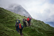 © anatoliy_gleb - Back view of tourists with backpacks using trekking poles while climbing the grassy hill. Group of active people walking on path and heading to foggy mountain. Concept of hiking and backpacking.