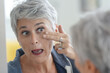 © goodluz - Mature white-haired woman checking eye wrinkles in front of mirror