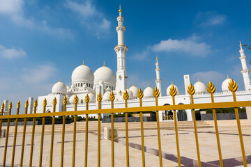 Wall Mural - Golden fence and blurred background of the white Grand Mosque built with marble stone against blue sky, also called Sheikh Zayed Grand Mosque in Abu Dhabi, UAE