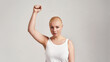 © Svitlana - Portrait of attractive young caucasian woman with shaved head wearing white shirt, looking at camera while raising clenched fist, standing isolated over grey background