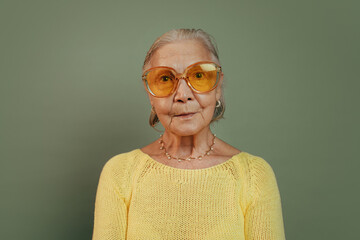 Wall Mural - Intelligent neutral old woman in cool orange sunglasses crossing her hands and looking at camera. Travel conception. Isolated over dark green background. Yellow casual clothes.