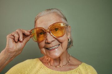 Wall Mural - Cheerful happy old lady smiles broadly showing her teeth and fixes big orange glasses on her face. Joyful elderly woman in yellow knitted sweater, necklace with big pearls and shiny earrings.