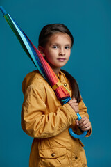 Wall Mural - portrait of little child girl with multicolored umbrella in yellow rain coat on blue background. autumn mood