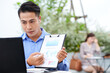 © DragonImages - Young Asian businessman working at outdoor cafe, video calling colleague and showing report