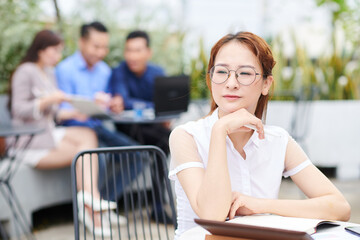 Wall Mural - Portrait of pensive young Vietnamese businesswoman sitting at table in outdoor cafe with opened planner and looking away