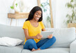 © Prostock-studio - Full length portrait of happy black woman using tablet computer on couch in living room