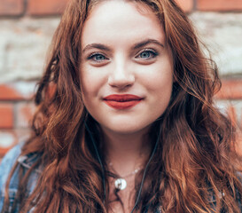 Wall Mural -  Smiling Red curled hair caucasian teen girl with applied red lipstick lips with blue eyes fashion vertical portrait on the red brick wall background. Natural people beauty concept image.