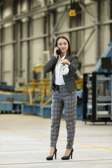 Wall Mural - Portrait of a female factory manager in a business suit holding mobile phone and retro alarm clock , controlling the work process in the helicopter manufacturer. Timeline. Deadline. 