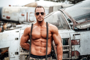 Wall Mural - Handsome muscular guy with naked torso and sunglasses in camouflage shorts poses near abandoned airplane in daytime.