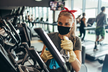 Young female worker disinfecting cleaning and weeping expensive fitness gym equipment with alcohol sprayer and cloth. Coronavirus global world pandemic and health protection safety measures.