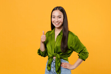 Wall Mural - Smiling cheerful beautiful attractive young brunette asian woman 20s wearing basic green shirt standing showing thumb up looking camera isolated on bright yellow colour background, studio portrait.