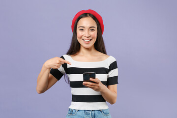Wall Mural - Smiling funny young asian woman 20s wearing striped t-shirt red beret pointing index finger on mobile cell phone typing sms message looking camera isolated on pastel violet background studio portrait.
