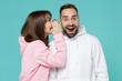 © ViDi Studio - Excited young couple two friends man woman 20s wearing white pink casual hoodie standing whispering secret behind hand, sharing news isolated on blue turquoise colour wall background studio portrait.