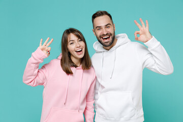 Cheerful excited young couple two friends man woman 20s wearing white pink casual hoodie standing showing OK gesture looking camera isolated on blue turquoise colour wall background studio portrait.