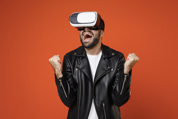 Wall Mural - Happy screaming young bearded man 20s wearing casual basic white t-shirt black leather jacket standing watching in vr headset gadget doing winner gesture isolated on orange background studio portrait.