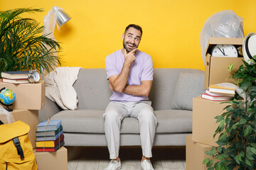 Wall Mural - Pensive owner man put hand prop up on chin sits in living room on sofa at home household unpacking stuff in polythene indoor rents flat isolated on yellow wall. Relocation moving in apartment concept.