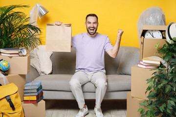 Wall Mural - Happy owner man doing winner gesture hold paper bag takeaway sit in living room on sofa at home household unpacking stuff rent flat isolated on yellow wall. Relocation moving in new apartment concept.