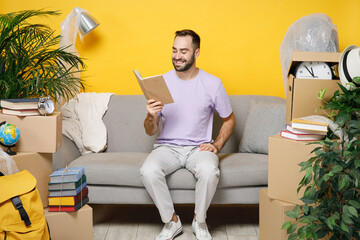 Wall Mural - Smiling cheerful young owner man reading book sits in living room on sofa at home household unpacking stuff indoor plant rents flat isolated on yellow wall. Relocation moving in new apartment concept.