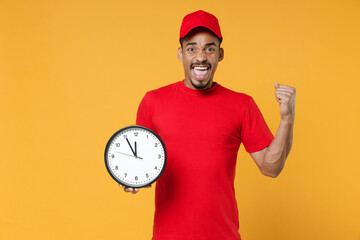 Delivery employee african man in red cap blank print t-shirt uniform workwear work courier service during quarantine coronavirus covid-19 virus concept hold clock isolated on yellow background studio.