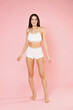 © ViDi Studio - Full length of smiling cheerful beautiful young brunette woman in white underwear showing fit sports body posing standing looking camera isolated on pastel pink colour background, studio portrait.