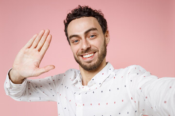 Wall Mural - Close up smiling young bearded man 20s in basic casual white shirt doing selfie shot on mobile phone waving greeting with hand looking camera isolated on pastel pink color background studio portrait.