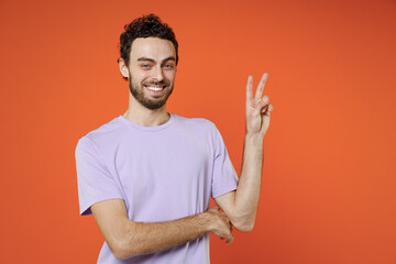 Wall Mural - Smiling funny cheerful handsome young bearded man 20s wearing basic casual violet t-shirt standing showing victory sign looking camera isolated on bright orange color wall background studio portrait.