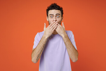 Wall Mural - Shocked worried scared young bearded man 20s wearing basic casual violet t-shirt standing covering mouth with hands looking camera isolated on bright orange color wall background studio portrait.