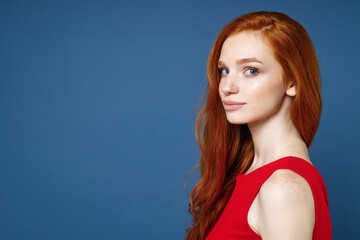 Wall Mural - Side view of smiling charming beautiful pretty young redhead woman 20s wearing bright red elegant evening dress standing and looking camera isolated on blue color wall background studio portrait.