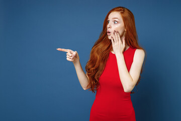 Wall Mural - Shocked young redhead woman 20s wearing bright red elegant evening dress standing pointing index finger aside on mock up copy space put hand on cheek isolated on blue color background studio portrait.