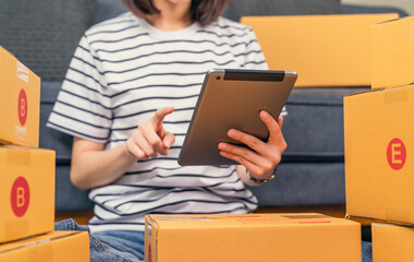 Wall Mural - Startup small business, A young Asian woman checking online order on digital laptop and packing boxes for products to send to customers. working at the home office.