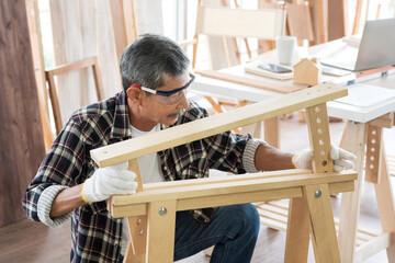 Senior asian old man carpenter wearing glasses and gloves protection building diy furniture in workshop