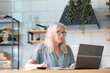 © yavdat - Senior woman using laptop for websurfing in her kitchen. The concept of senior employment, social security. Mature lady sitting at work typing a notebook computer in an home office.