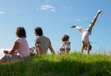 Back View Of A Couple With Children Sitting On A Hill Against Blue Sky And Girl Doing A Handstand