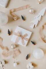 Poster - Paper gift box with bow tie and Christmas toys: fir-trees, baubles, ribbons on pastel beige background. Holiday celebration concept. Top view, flat lay. Christmas / New Year decoration.