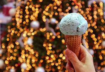 Wall Mural - Closeup a Scoop of Mint Chocolate Chip Ice Cream Cone in Hand with Glittering Christmas Tree Bokeh in Background