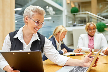 portrait of adult business lady working on laptop in the office with colleagues in the background, focus on short-haired female in formal wear at table