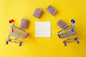 Two mini shopping cart with small craft bags and blank white paper for lettering or shopping list on yellow background.