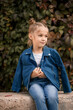 © Дмитрий Ткачук - A beautiful stylish blonde girl in a blue jacket sits on a concrete block. Girl 7 years old with a smile on a background of leaves