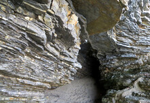 Rock With Geological Layers Of Gray And Yellow Stone. Sandy Path Entrance To The Cave In The Mountain.