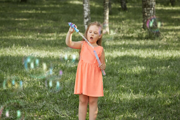 A little girl in an orange dress on summer vacation blows soap bubbles. Sunny summer warm day.