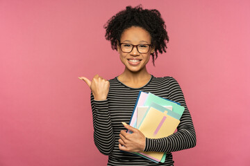 Afro-American tutor or teacher woman isolated on pink studio wall. Student girl wear glasses holding notebooks, smiling, showing finger on free space. Education in high school university college.
